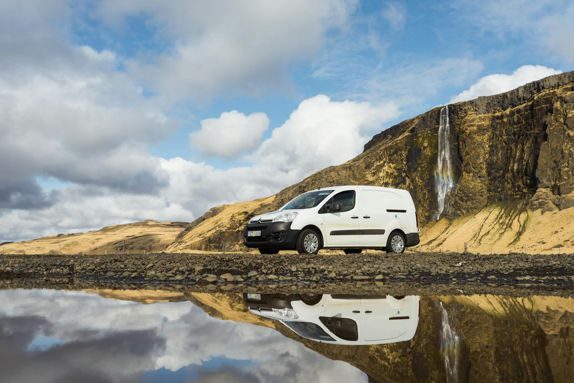 Une camionnette Citroen Berlingo garée par un lac réfléchissant avec des montagnes et une cascade en arrière-plan.