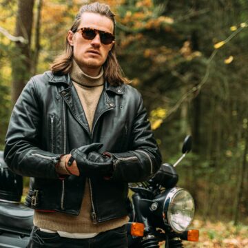 Motard confiant en veste de cuir noir debout en moto dans un cadre forestier.